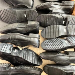 May include: A collection of black leather dress shoes, displayed with their soles facing upwards. The shoes have a classic design with laces and rubber soles. The image shows a variety of sizes and styles.