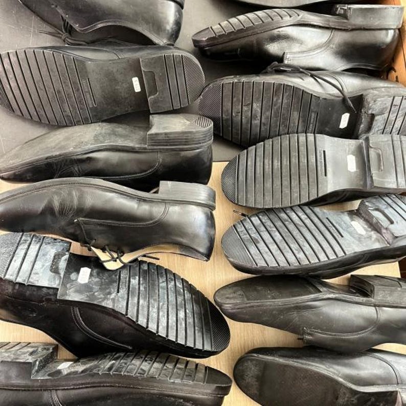 May include: A collection of black leather dress shoes, displayed with their soles facing upwards. The shoes have a classic design with laces and rubber soles. The image shows a variety of sizes and styles.