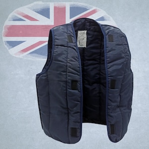 May include: A navy blue quilted vest with a dark blue lining. The sleeveless vest features a front closure with black velcro patches. A tag is visible inside. The background shows a faded Union Jack.