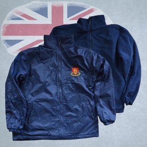 Op de afbeelding: Twee marineblauwe jassen worden getoond tegen een vervaagde Union Jack-achtergrond. Eén is een windjack met een rits en een embleem op de borst. De andere is een fleecejack met een hoge kraag. Beide jassen zijn effen marineblauw.