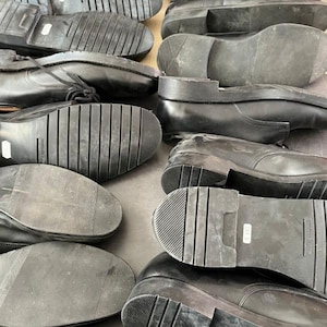 May include: A collection of black leather shoes, viewed from the soles. The soles have various tread patterns, including ribbed and textured designs. Some shoes have laces, while others are slip-on styles. The shoes appear to be used.