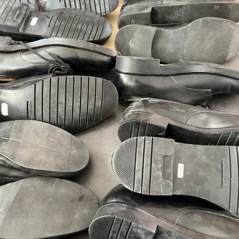 May include: A collection of black leather shoes, viewed from the soles. The soles have various tread patterns, including ribbed and textured designs. Some shoes have laces, while others are slip-on styles. The shoes appear to be used.