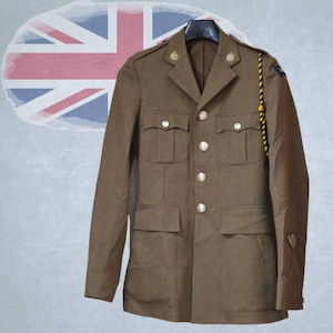Op de afbeelding: Een beige jas in militaire stijl met een ingekeepte revers, vier voorzakken met knoopsluitingen en een centrale rij zilverkleurige knopen. Een zwart-geel gevlochten koord hangt aan de linkerschouder en kleine emblemen zijn zichtbaar.