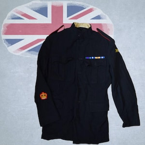 May include: A dark navy blue military-style jacket featuring a high collar, two chest pockets, and shoulder epaulets with thin red piping. The jacket displays a red crown patch on the right sleeve, a yellow emblem on the left, and a colorful ribbon bar.