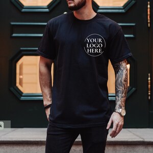 May include: Black t-shirt with a round white logo that says "YOUR LOGO HERE". The shirt is short-sleeved and appears to be made of a soft material. The person wearing the shirt has a tattoo on their arm.