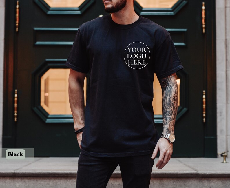 May include: Black t-shirt with a round white logo that says "YOUR LOGO HERE". The shirt is short-sleeved and appears to be made of a soft material. The person wearing the shirt has a tattoo on their arm.