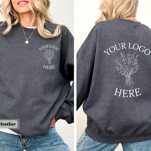 May include: Dark heather gray sweatshirt with white text and a floral design. The front of the sweatshirt has the text "YOUR LOGO HERE" and a floral design. The back of the sweatshirt has the same text and design. The bottom left corner of the image has the text "Dark Heather".