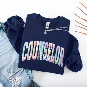 Peut inclure: Un sweat-shirt bleu marine avec le mot "COUNSELOR" en lettres patchwork color&eacute;es. Un collier argent&eacute; avec un petit pendentif est pos&eacute; sur le sweat-shirt. Le sweat-shirt est &agrave; c&ocirc;t&eacute; d'un jean bleu d&eacute;chir&eacute;.