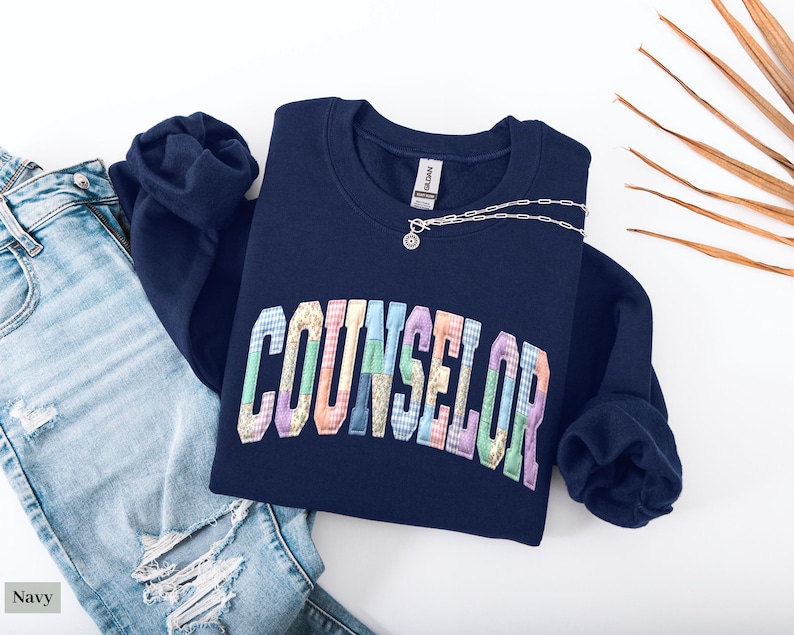 Peut inclure: Un sweat-shirt bleu marine avec le mot "COUNSELOR" en lettres patchwork color&eacute;es. Un collier argent&eacute; avec un petit pendentif est pos&eacute; sur le sweat-shirt. Le sweat-shirt est &agrave; c&ocirc;t&eacute; d'un jean bleu d&eacute;chir&eacute;.