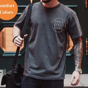 May include: A dark gray t-shirt with a round logo that says "YOUR LOGO HERE". The shirt is worn with a black shoulder bag. The image also includes the text "Comfort Colors" in an orange circle and the word "Pepper" in a gray rectangle.