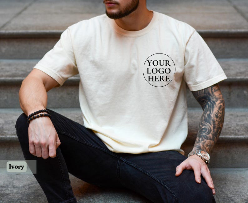 May include: Ivory-colored t-shirt with a crew neck and short sleeves. The shirt features a circular logo design that says "YOUR LOGO HERE". The model is wearing black jeans and a watch. The shirt is a neutral color.