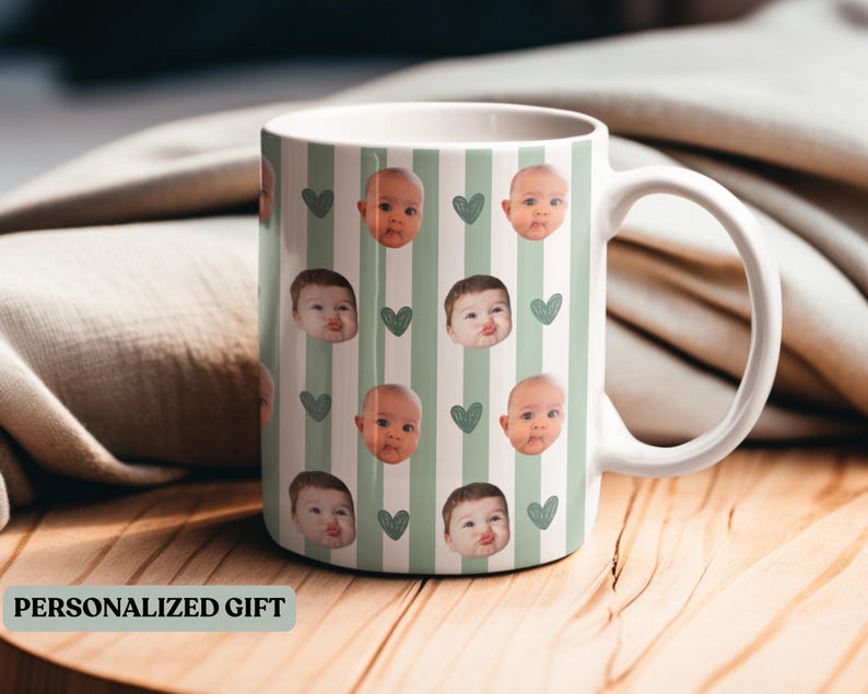 May include: White ceramic mug featuring a pattern of baby faces and green hearts on a striped background. The mug has a white handle and the text "PERSONALIZED GIFT" is visible.