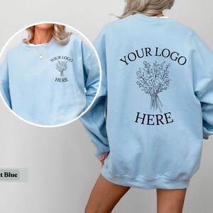 May include: Light blue sweatshirt with the text "YOUR LOGO HERE" and a floral bouquet design on the back. The front of the sweatshirt also features the text "YOUR LOGO HERE" and a floral bouquet design. The sweatshirt is a light blue color.