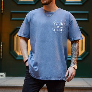 May include: A blue jean-colored, short-sleeved t-shirt with a crew neck. The shirt has a faded appearance and features a circular logo design that reads "YOUR LOGO HERE". The shirt is worn with black pants.