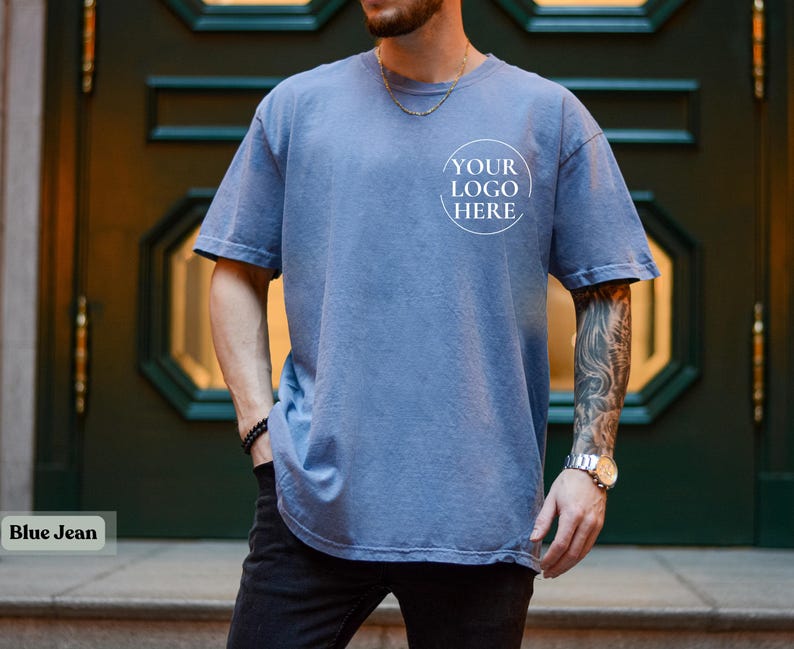 May include: A blue jean-colored, short-sleeved t-shirt with a crew neck. The shirt has a faded appearance and features a circular logo design that reads "YOUR LOGO HERE". The shirt is worn with black pants.