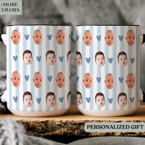 May include: Two white ceramic mugs with black handles and rims. The mugs display a pattern of baby faces, blue hearts, and vertical blue stripes. The text "+MORE COLORS" and "PERSONALIZED GIFT" are visible.