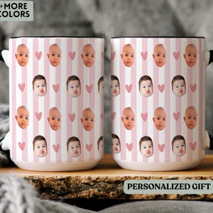 May include: Two white ceramic mugs with black handles. The mugs have a repeating pattern of baby faces, pink hearts, and pink and white stripes. The text "PERSONALIZED GIFT" is visible on the bottom of the mug.