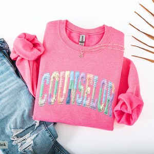 Peut inclure: Un sweat-shirt rose vif avec le mot "COUNSELOR" en lettres patchwork color&eacute;es. Il est associ&eacute; &agrave; un jean bleu clair d&eacute;chir&eacute; et un collier argent&eacute;. Les mots "Safety Pink" sont dans le coin inf&eacute;rieur gauche.