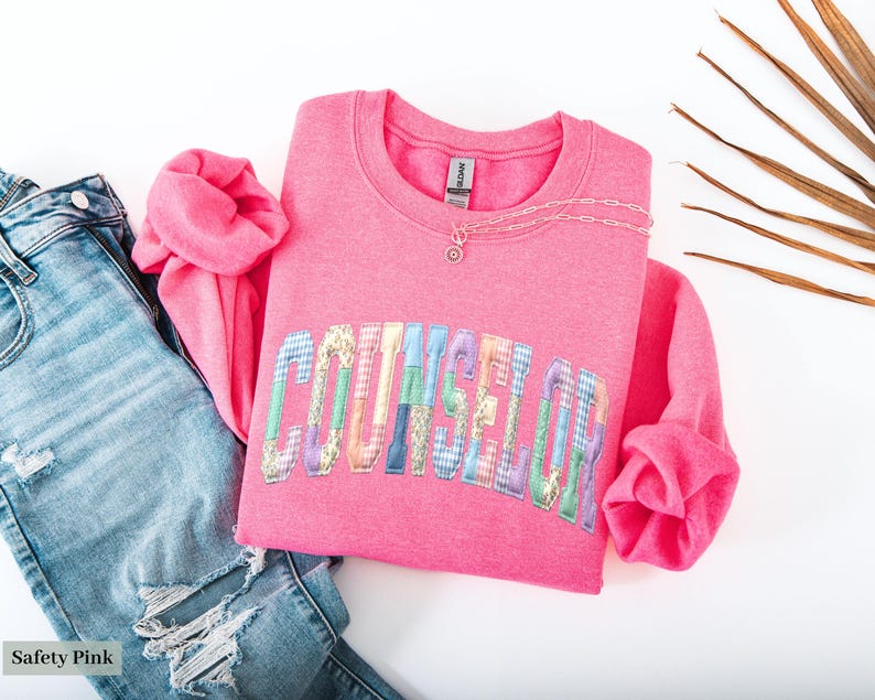Peut inclure: Un sweat-shirt rose vif avec le mot "COUNSELOR" en lettres patchwork color&eacute;es. Il est associ&eacute; &agrave; un jean bleu clair d&eacute;chir&eacute; et un collier argent&eacute;. Les mots "Safety Pink" sont dans le coin inf&eacute;rieur gauche.