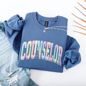 Peut inclure: Sweat-shirt bleu indigo avec le mot "COUNSELOR" en lettres patchwork color&eacute;es. Le sweat-shirt est associ&eacute; &agrave; un jean d&eacute;chir&eacute; bleu clair et &agrave; un collier argent&eacute;. Une feuille de palmier s&eacute;ch&eacute;e est en arri&egrave;re-plan.