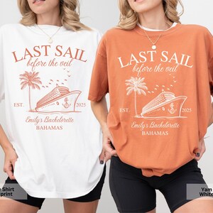 May include: Two white t-shirts with a peach colored print. The print features a cruise ship, palm tree, and the text "Last Sail Before the Veil", "Est. 2025", "Emily's Bachelorette", and "Bahamas". One shirt is white with peach print and the other is peach with white print.