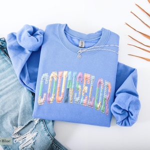 Peut inclure: Un sweat-shirt bleu clair avec le mot "COUNSELOR" en lettres patchwork color&eacute;es. Un collier en argent avec un petit pendentif repose sur l'encolure. Le sweat-shirt est associ&eacute; &agrave; un jean en denim d&eacute;lav&eacute; clair avec des d&eacute;tails d&eacute;chir&eacute;s.