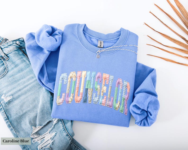 Peut inclure: Un sweat-shirt bleu clair avec le mot "COUNSELOR" en lettres patchwork color&eacute;es. Un collier en argent avec un petit pendentif repose sur l'encolure. Le sweat-shirt est associ&eacute; &agrave; un jean en denim d&eacute;lav&eacute; clair avec des d&eacute;tails d&eacute;chir&eacute;s.