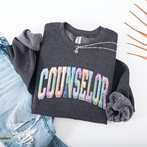 Peut inclure: Sweat-shirt gris fonc&eacute; avec le mot "COUNSELOR" en lettres multicolores. Un collier argent&eacute; avec un petit pendentif est pos&eacute; sur le sweat-shirt. Un jean bleu clair d&eacute;chir&eacute; est &agrave; c&ocirc;t&eacute;.
