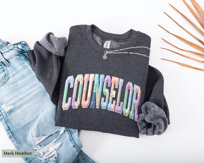 Peut inclure: Sweat-shirt gris fonc&eacute; avec le mot "COUNSELOR" en lettres multicolores. Un collier argent&eacute; avec un petit pendentif est pos&eacute; sur le sweat-shirt. Un jean bleu clair d&eacute;chir&eacute; est &agrave; c&ocirc;t&eacute;.