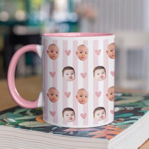 May include: A white ceramic mug with a pink interior and handle. The mug has a pattern of baby faces and pink hearts on a striped background. The mug is on a book with a floral design.