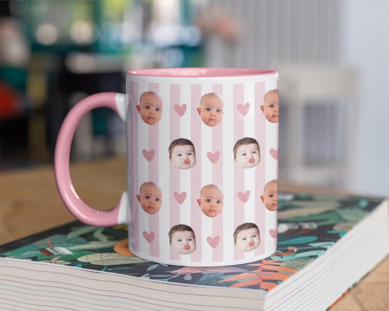 May include: A white ceramic mug with a pink interior and handle. The mug has a pattern of baby faces and pink hearts on a striped background. The mug is on a book with a floral design.