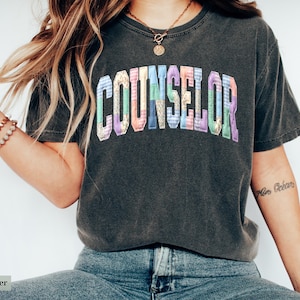 Peut inclure: T-shirt gris fonc&eacute; avec le mot "COUNSELOR" en lettres patchwork color&eacute;es. Le t-shirt a un aspect vintage et est fait d'une mati&egrave;re douce. Le design est centr&eacute; sur la poitrine.