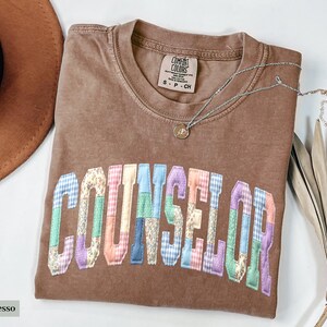 Peut inclure: Un t-shirt marron avec le mot "COUNSELOR" en lettres patchwork color&eacute;es. Le t-shirt a un col rond et une petite &eacute;tiquette. Un collier argent&eacute; avec un pendentif est drap&eacute; sur le t-shirt.
