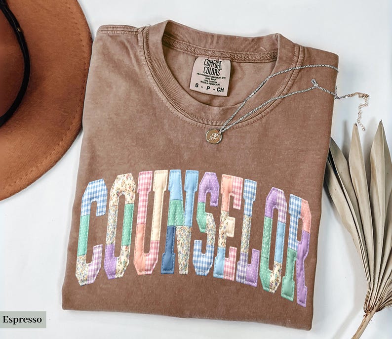 Peut inclure: Un t-shirt marron avec le mot "COUNSELOR" en lettres patchwork color&eacute;es. Le t-shirt a un col rond et une petite &eacute;tiquette. Un collier argent&eacute; avec un pendentif est drap&eacute; sur le t-shirt.