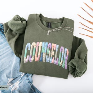 Peut inclure: Un sweat-shirt vert olive avec le mot "COUNSELOR" dans un design patchwork color&eacute;. Un collier en argent avec un petit pendentif est drap&eacute; sur le sweat-shirt. Le sweat-shirt est associ&eacute; &agrave; un jean bleu clair d&eacute;chir&eacute;.