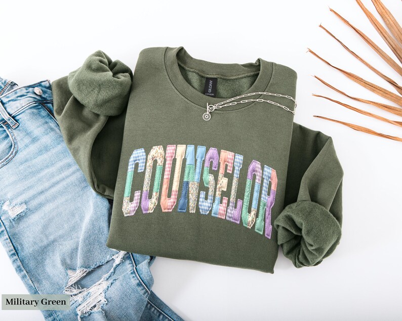 Peut inclure: Un sweat-shirt vert olive avec le mot "COUNSELOR" dans un design patchwork color&eacute;. Un collier en argent avec un petit pendentif est drap&eacute; sur le sweat-shirt. Le sweat-shirt est associ&eacute; &agrave; un jean bleu clair d&eacute;chir&eacute;.