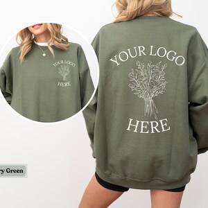 May include: Military green sweatshirt with a crew neck. The front features a small graphic and the text "YOUR LOGO HERE". The back displays a larger floral design and the same text. The sweatshirt is a solid color.