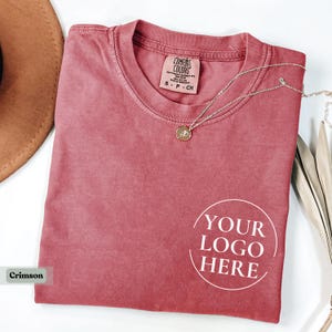 May include: Crimson-colored t-shirt with the text "YOUR LOGO HERE" in a white circle. A silver necklace with a pendant rests on the shirt. The shirt is folded and placed on a white surface.