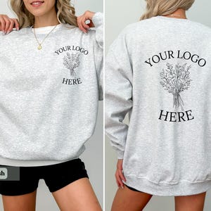 May include: Light gray sweatshirt with the text "YOUR LOGO HERE" and a floral illustration on the front and back. The sweatshirt has a crew neck and long sleeves. The bottom of the sweatshirt is white.