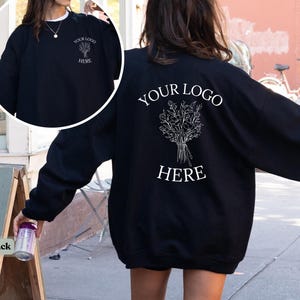 May include: Black sweatshirt with white text and a floral design. The front of the sweatshirt has a small floral design and the text "YOUR LOGO HERE". The back of the sweatshirt has a larger floral design and the text "YOUR LOGO HERE".