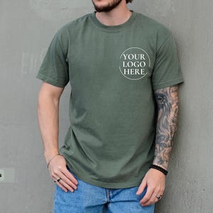 May include: Moss green t-shirt with a crew neck and short sleeves. The shirt features a circular logo that says "YOUR LOGO HERE" on the front. The model is wearing blue jeans.