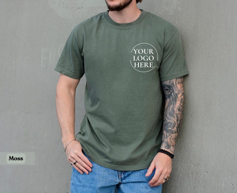 May include: Moss green t-shirt with a crew neck and short sleeves. The shirt features a circular logo that says "YOUR LOGO HERE" on the front. The model is wearing blue jeans.