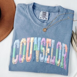 Peut inclure: Un t-shirt bleu Comfort Colors avec le mot "COUNSELOR" en lettres patchwork color&eacute;es. Un collier en or avec un pendentif repose sur le t-shirt. Le t-shirt est pli&eacute;, et un chapeau marron est visible en arri&egrave;re-plan.