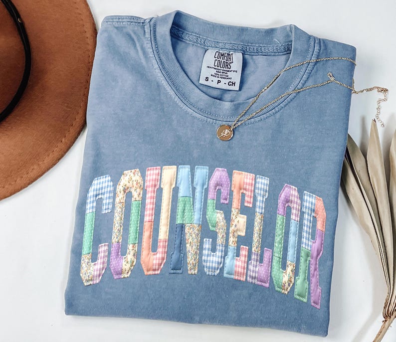 Peut inclure: Un t-shirt bleu Comfort Colors avec le mot "COUNSELOR" en lettres patchwork color&eacute;es. Un collier en or avec un pendentif repose sur le t-shirt. Le t-shirt est pli&eacute;, et un chapeau marron est visible en arri&egrave;re-plan.