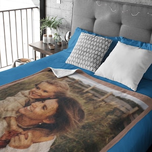 May include: A personalized blanket featuring a photograph of a woman and child embracing, set against a grassy background. The blanket has a brown border and is displayed on a blue bed with decorative pillows.