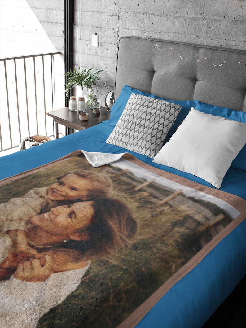 May include: A personalized blanket featuring a photograph of a woman and child embracing, set against a grassy background. The blanket has a brown border and is displayed on a blue bed with decorative pillows.