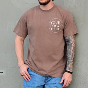 May include: Espresso-colored short-sleeve t-shirt with a crew neck. The shirt features a white circular logo that says "YOUR LOGO HERE". The model is wearing blue jeans. The shirt is a solid color.