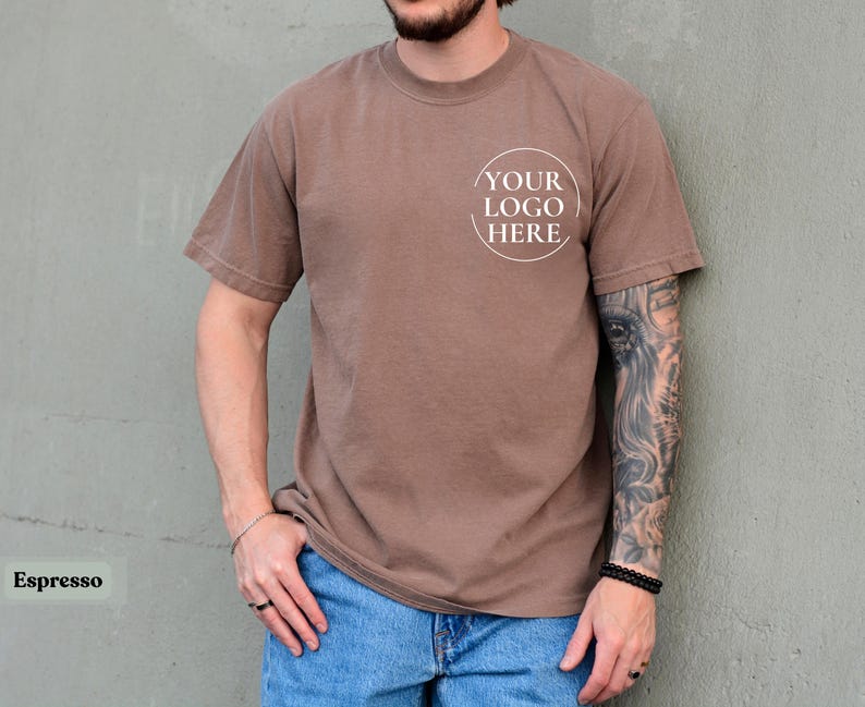 May include: Espresso-colored short-sleeve t-shirt with a crew neck. The shirt features a white circular logo that says "YOUR LOGO HERE". The model is wearing blue jeans. The shirt is a solid color.