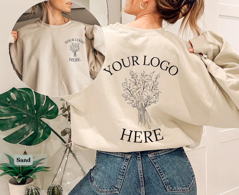 May include: A sand-colored sweatshirt with the text "YOUR LOGO HERE" and a floral bouquet design on the front and back. The sweatshirt is a crew neck style and is made of a soft material. The model is wearing blue jeans.
