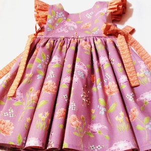 May include: A lilac-colored dress with a floral pattern featuring peach and pink flowers, green leaves, and small white blossoms. The dress has ruffled sleeves and peach-colored bows at the shoulders. The dress is designed for a child.